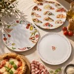 Tre piatti da pizza decorati disposti su una tovaglia chiara accanto a pomodori freschi e una pizza margherita fumante