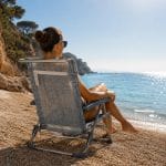 Donna seduta su una sedia pieghevole da spiaggia azzurra con braccioli e schienale alto vista mare